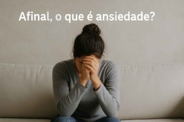 A imagem mostra uma mulher jovem, de pele morena clara e cabelos escuros presos em um coque, sentada sozinha em um sofá bege claro. Ela está curvada para frente, com os cotovelos apoiados nos joelhos e o rosto encostado nas mãos, transmitindo um sentimento de cansaço emocional, tristeza ou ansiedade. A cena é minimalista, com fundo neutro e iluminação suave, o que reforça a atmosfera introspectiva e sensível. No topo da imagem, em letras brancas e grandes, aparece a pergunta: "Afinal, o que é ansiedade?"